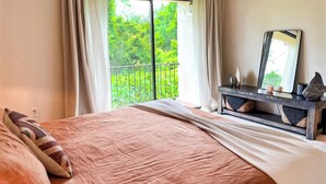 Ein Schlafzimmer mit einem großen Bett, einer Kommode mit Dekoration, einem Spiegel und Blick auf Grün durch ein Fenster.