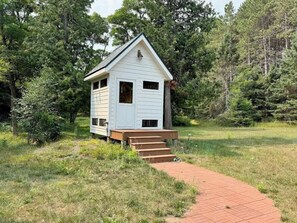 A small white cabin with a single door and two windows, surrounded by trees and a grassy area.