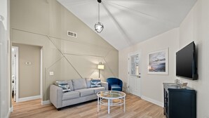 Een moderne woonkamer met een hoge plafond, een grijze bank, een ronde salontafel, een blauwe fauteuil, een zwarte kast en een tv aan de muur.