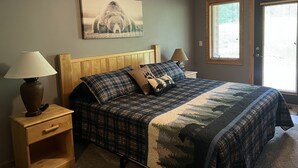 Ein Schlafzimmer mit einem Holzkopfteil, einem großen Bett mit karierter Bettdecke, zwei Nachttischen mit Lampen, einem Deckenventilator und einem Fenster mit Jalousien.
