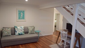 A living room with a grey sofa, teal cushions, a small round table, and a wooden chair. A staircase leads to another room. - Ashmore - Four bedroom home, easy walk to shops and beach, back deck with a view (South West Rocks)