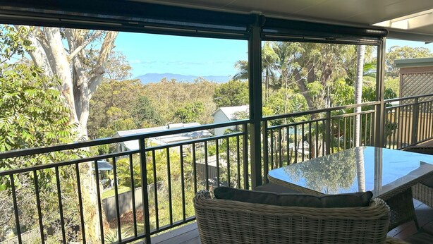 A balcony with a glass table and wicker chairs, overlooking a lush landscape with trees and a distant mountain range. - Ashmore - Four bedroom home, easy walk to shops and beach, back deck with a view (South West Rocks)