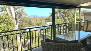 A balcony with a glass table and wicker chairs, overlooking a lush landscape with trees and a distant mountain range.