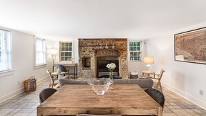 A dining area with a wooden table, cushioned chairs, a brick fireplace, and a large framed picture on the wall.