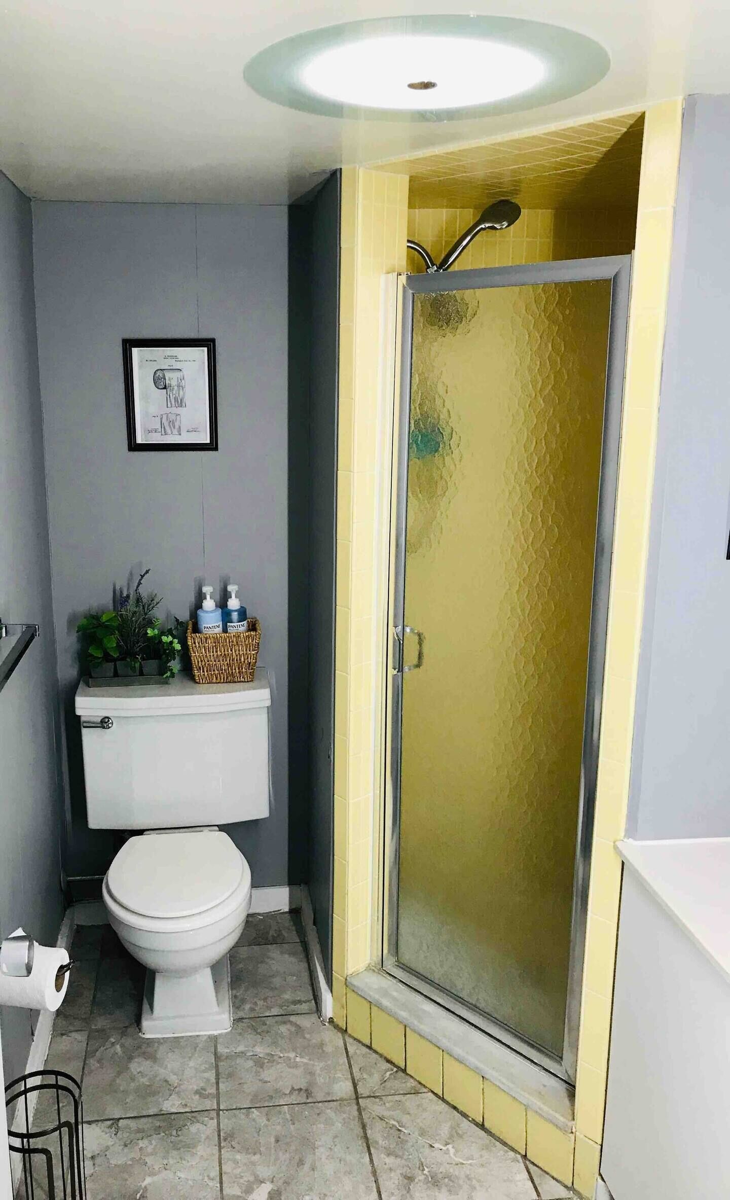 A bathroom with a toilet, a shower with a glass door, a framed certificate on the wall, and a basket with toiletries.