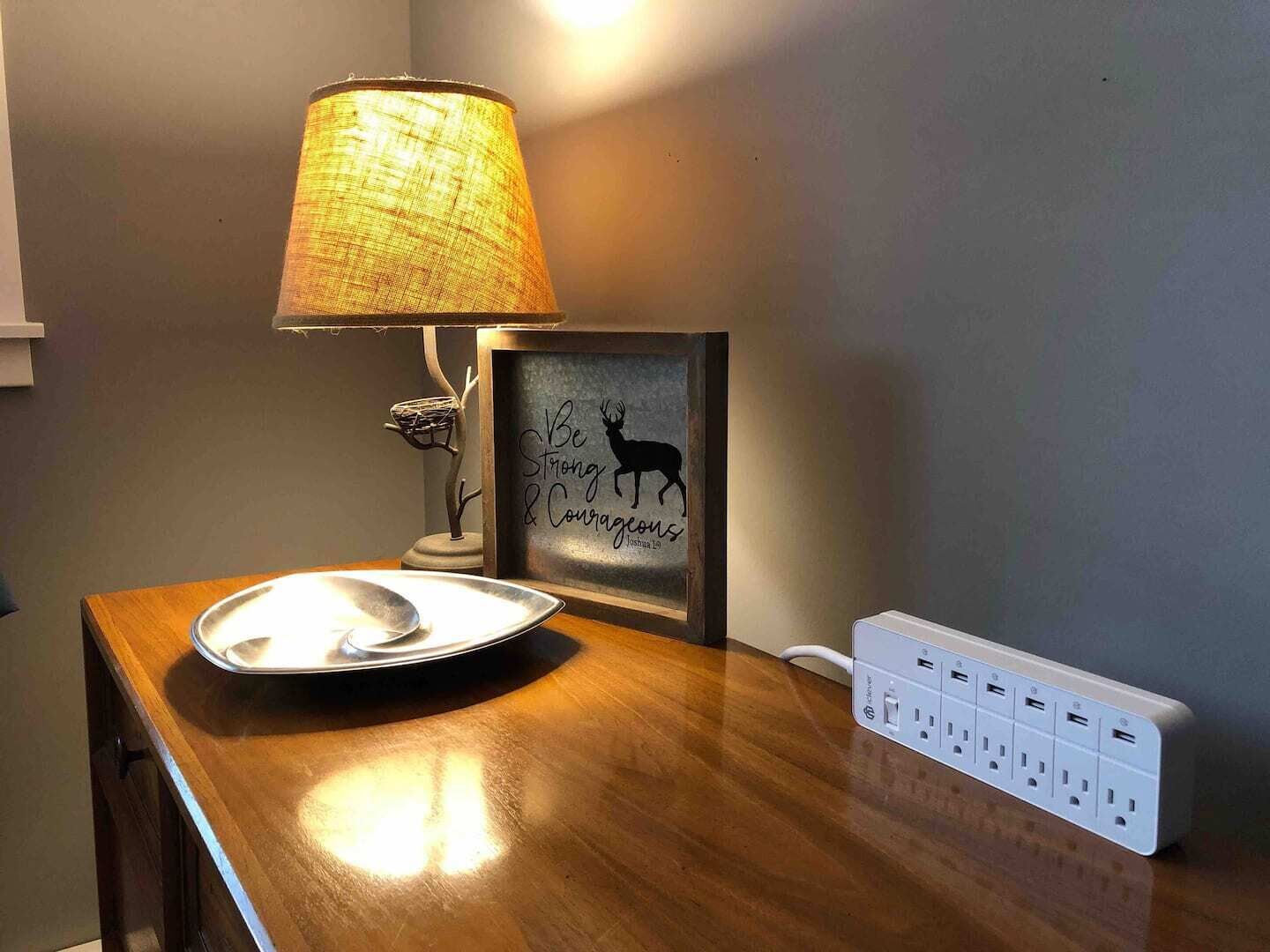 A wooden table with a metal tray, a lamp, a framed motivational quote, and a power strip.