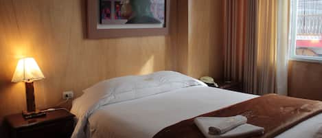 A hotel room with a bed, a nightstand with a lamp, a framed artwork on the wall, and a window with curtains.