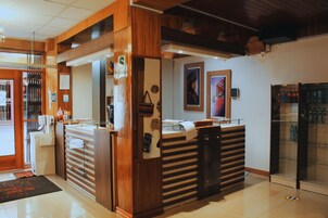 Un mostrador de recepción con detalles de madera, un separador de vidrio y una pantalla digital. En la pared hay obras de arte enmarcadas y un refrigerador con botellas.