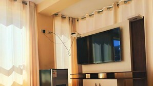 A living room with a flat-screen TV mounted on the wall, a built-in entertainment unit, a refrigerator, and a window with curtains. - KAARO HOTEL SUITE (Puno)