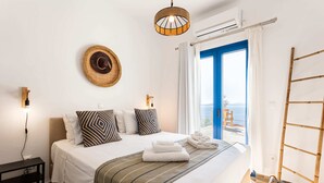 Un letto ben rifatto con lenzuola a righe, un cappello appeso al muro e vista sul mare attraverso una porta blu.