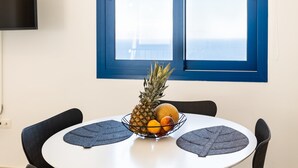 A dining table with a fruit bowl containing pineapple, melon, and orange. The table is round with a white surface and black chairs. There is a blue-framed window above the table. - Live In Blue - Uranian Private Pool & Magic Views (Coressia)