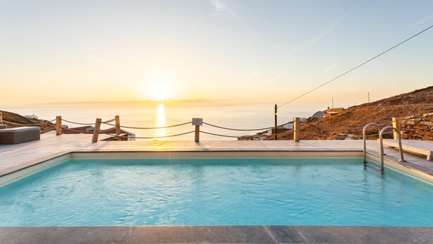 Une piscine avec vue sur le coucher de soleil sur la mer, entourée d’un mur de pierre et d’une terrasse en bois.