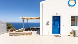 Un bâtiment blanc avec une porte bleue et une fenêtre ronde. On y trouve une pergola d’où l’on peut admirer la mer.