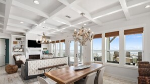 Uma sala de estar ampla com uma mesa de jantar de madeira, assentos confortáveis, lareira e grandes janelas com vista para o oceano.