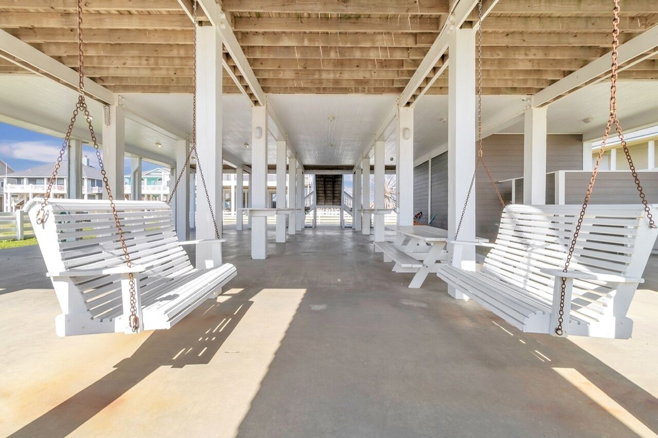 A covered outdoor area with white benches and picnic tables, supported by white columns, under a wooden ceiling.
