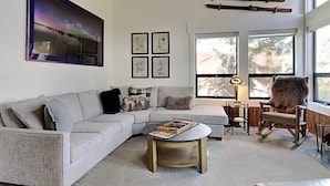 A modern living room with a grey sectional sofa, a coffee table, and a rocking chair. There are framed artworks on the wall and a large window providing natural light. - Summer Fun - SLOPESIDE 4BR/3BA with 3 Spas, Pool and Sauna - Perfect for Family and Friend Getaways All Year Round by RedAwning (Mammoth Lakes)