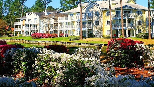 Um hotel com vários andares, fachada simétrica, varandas e um jardim bem cuidado, com arbustos e flores diversas.