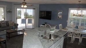 A modern kitchen with a large island, a dining area with white chairs, a sofa with decorative pillows, and a ceiling fan.