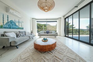 Ein geräumiges Wohnzimmer mit großem Fenster, einem modernen Kronleuchter und einer Sofagarnitur.