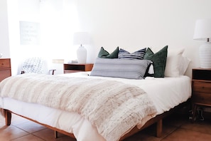 Una cama bien tendida con una colcha blanca y una manta decorativa estampada. Hay varios cojines de diferentes colores y diseños. Se ve una mesita de noche con una lámpara y un objeto decorativo pequeño. También hay una estantería con libros y una cita enmarcada.