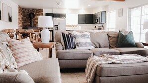 Een moderne woonkamer met een bank, fauteuils en een salontafel. Er is een open haard, een keuken op de achtergrond en grote ramen.