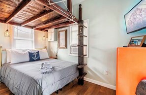 A bedroom with a bed, wooden beams, a ladder, a television, and a window.