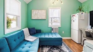 Une pièce avec un canapé bleu, un réfrigérateur blanc, un luminaire au plafond et une fenêtre avec des stores.
