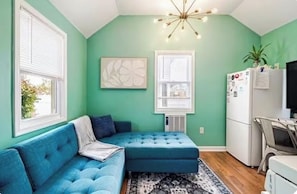 A room with a blue sofa, a white refrigerator, a ceiling light fixture, and a window with blinds.