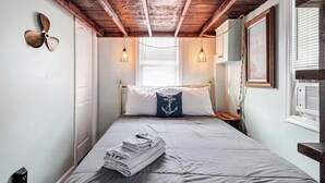 Une chambre avec un plafond en bois, un lit avec un oreiller bleu, un ventilateur fixé au mur et une fenêtre avec des stores.