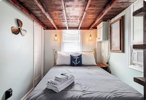 A bedroom with a wooden ceiling, a bed with a blue pillow, a fan on the wall, and a window with blinds.