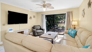 A living room with a beige sofa, a leather armchair, a coffee table, a flat-screen TV, a ceiling fan, and a sliding glass door leading to an outdoor patio. - Charming Pet Friendly Villa - Steps to the Beach and Coligny Plaza! (Hilton Head Island)