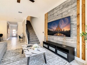 A living room with a large flat-screen TV mounted on a wooden wall, a staircase, a dining area, and a sofa.