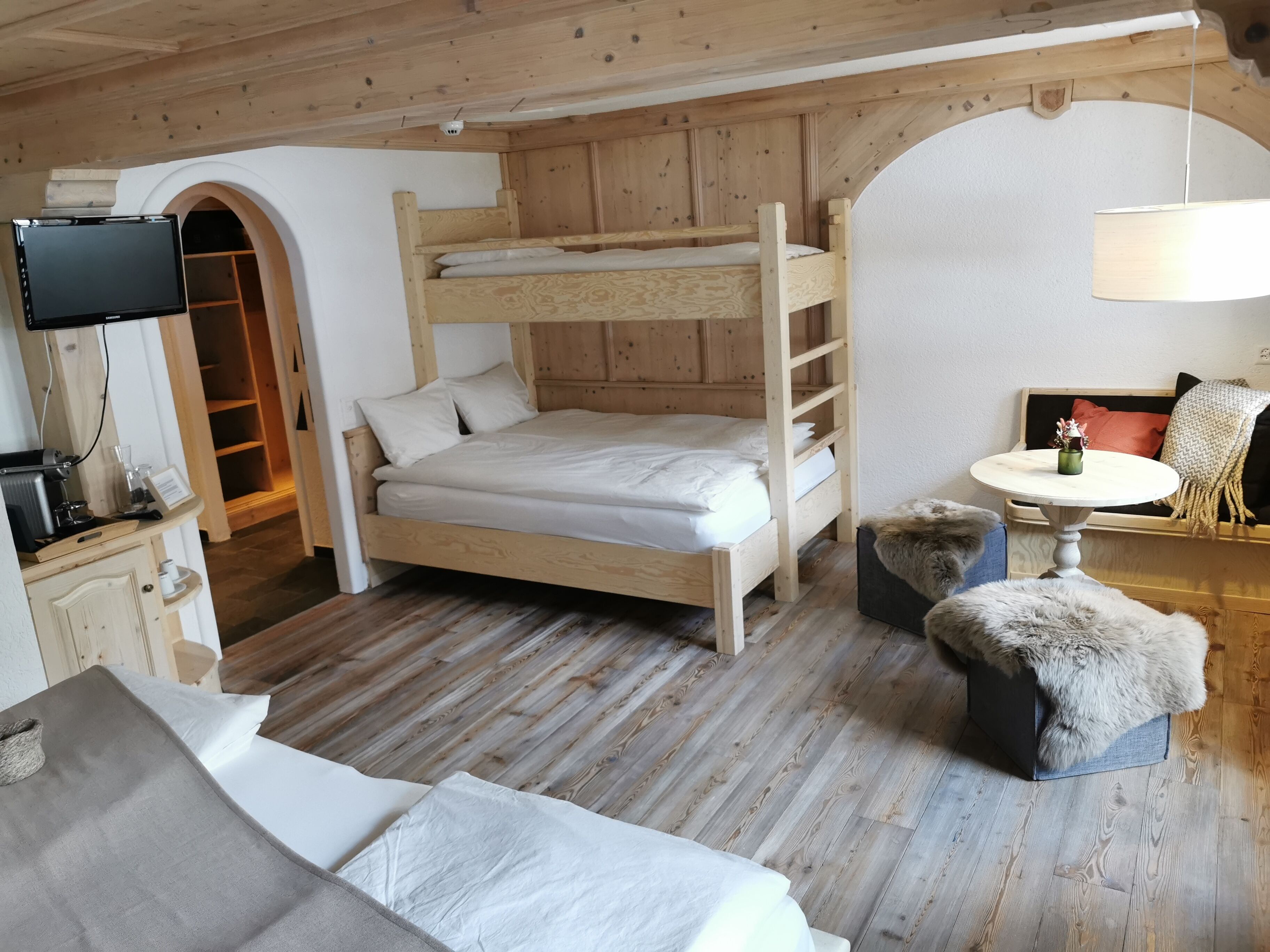 A cozy room with a bunk bed, a small table with a lamp, a sofa, and a TV mounted on the wall.