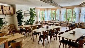 A spacious dining area with wooden tables and chairs, large windows with sheer curtains, and potted plants.