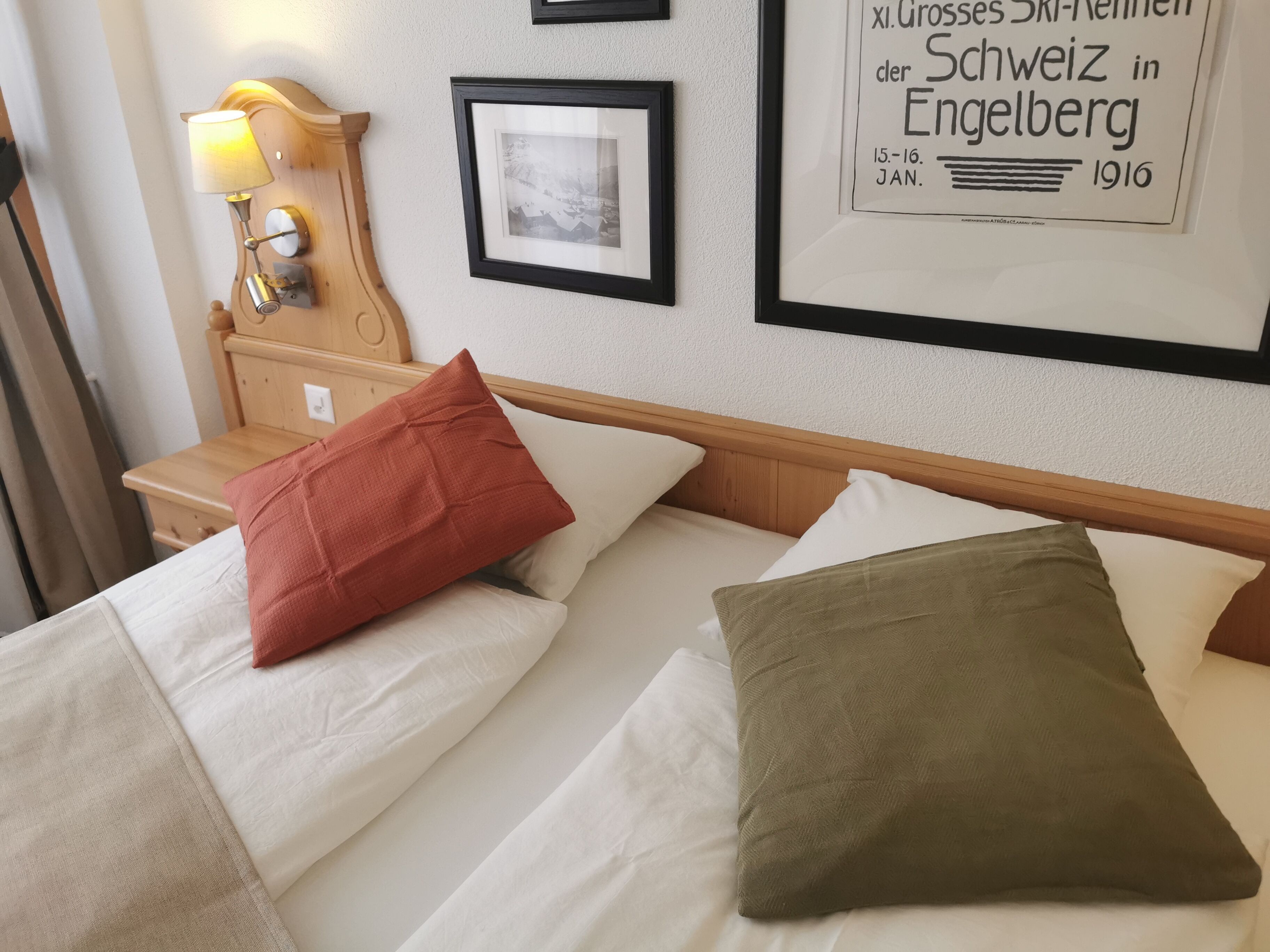 Een hotelkamer met een houten hoofdbord, twee bedden met witte lakens, een rood en een groen kussen, een nachtkastje met een lamp, en een ingelijst schilderij aan de muur.