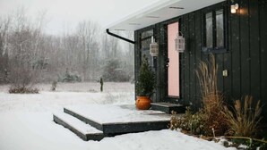 Exterior nevada d'una casa moderna amb façana de fusta fosca, una porta visible i una petita zona de porxo amb escales que hi condueixen.