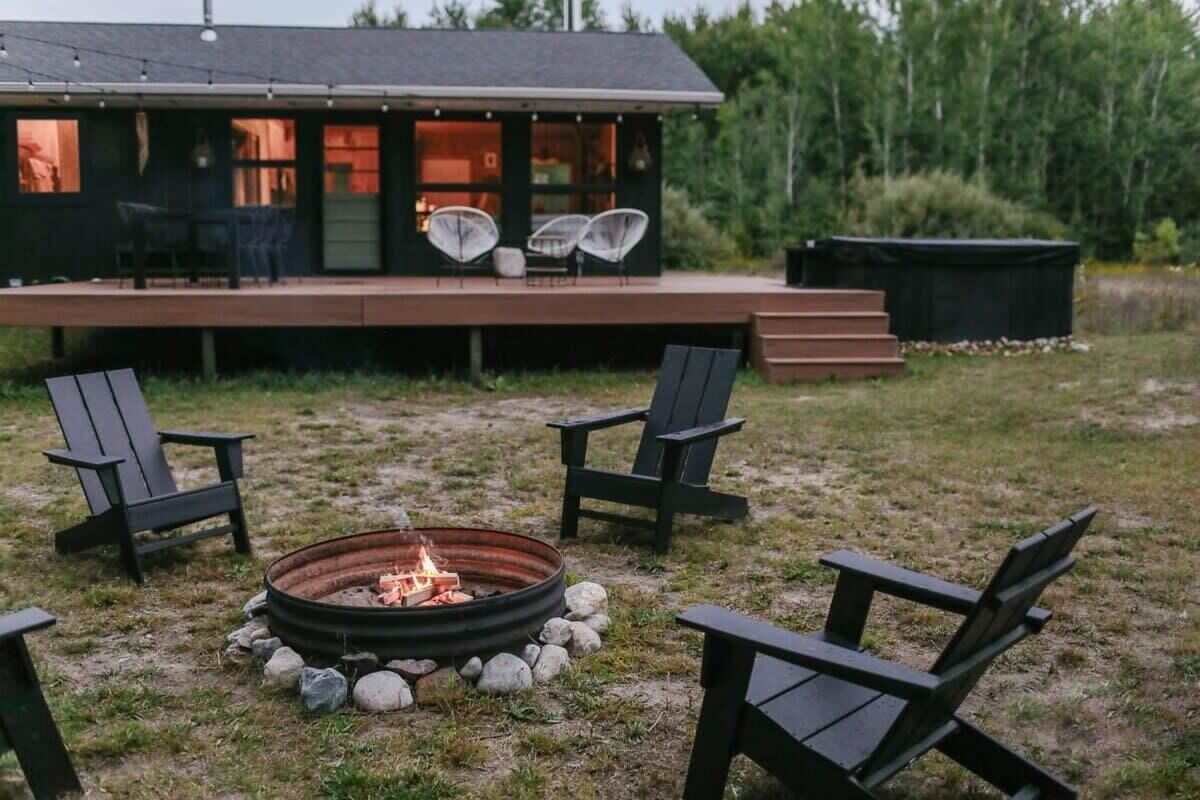 Een gezellig buitenverblijf met een vuurkuil, omringd door Adirondack-stoelen, voor een modern huis met een terras en grote ramen.
