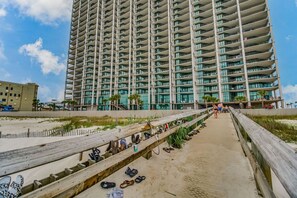 A high-rise building with multiple balconies, a wooden boardwalk with shoes, and a beach in the background. - Gulf Front - 3BR 4BA - Lazy River - The Oasis at Orange Beach (Orange Beach)