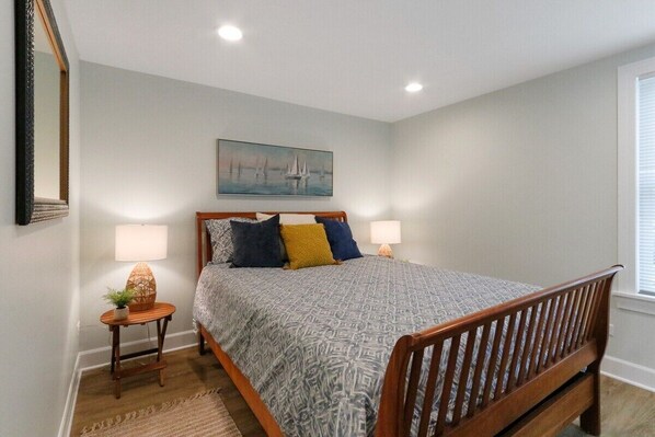 A bedroom with a bed, two bedside lamps, a nightstand with a plant, a painting on the wall, and a window with blinds.