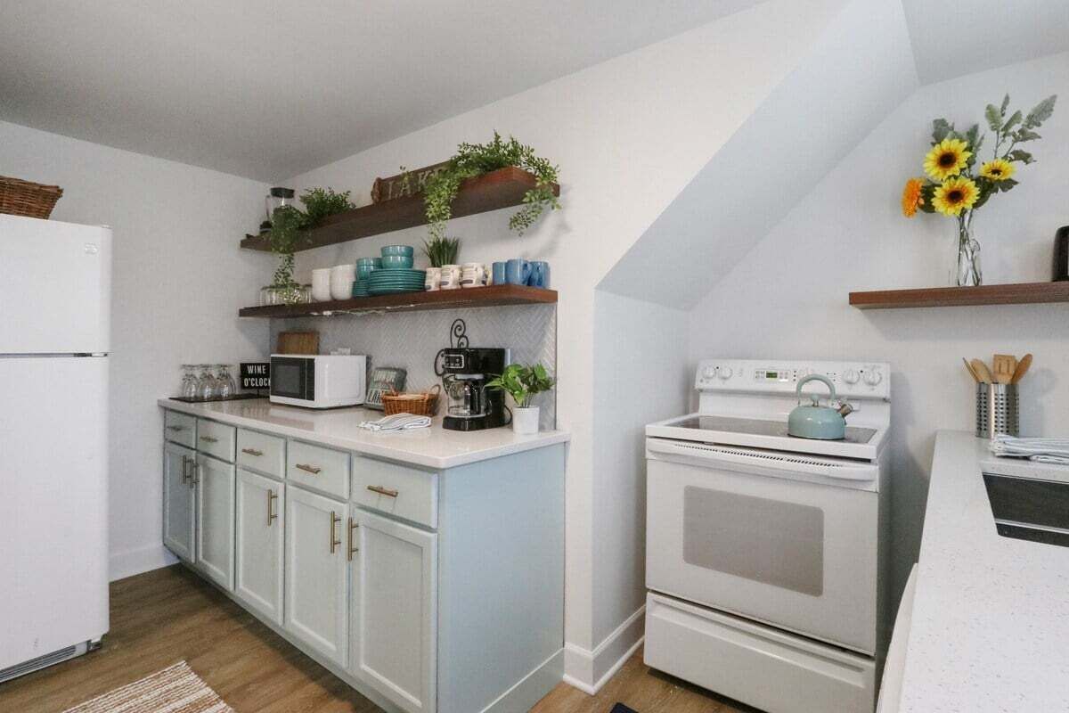 Eine Küche mit weißen Schränken, einem weißen Kühlschrank, einem weißen Herd und einer weißen Arbeitsplatte. Regale mit Pflanzen und Geschirr sind ebenfalls vorhanden.