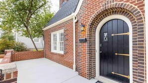 Uma casa de tijolos com porta preta, persianas brancas e uma pequena varanda.