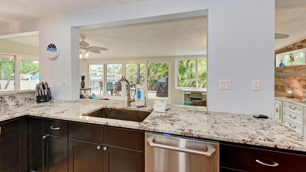 A kitchen with dark cabinets, a stainless steel dishwasher, a granite countertop, and a large window overlooking greenery. - Salty Sea Life - Canal Pool Home-Just 5 Min to Anna Maria Island - By Island Rea (Bradenton)