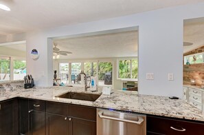 A kitchen with dark cabinets, a stainless steel dishwasher, a granite countertop, and a large window overlooking greenery.