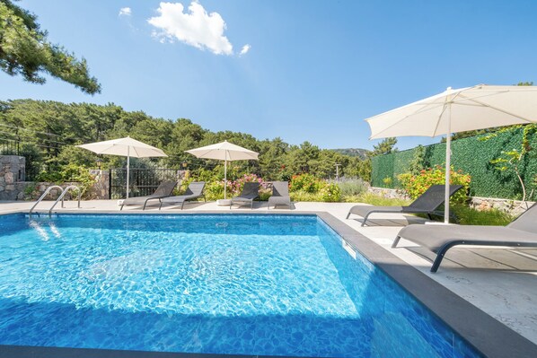 Une piscine entourée de verdure et d’un mur de pierre, avec des chaises longues et des parasols.