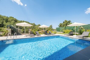 A swimming pool with lounge chairs and umbrellas, surrounded by greenery and a clear sky.