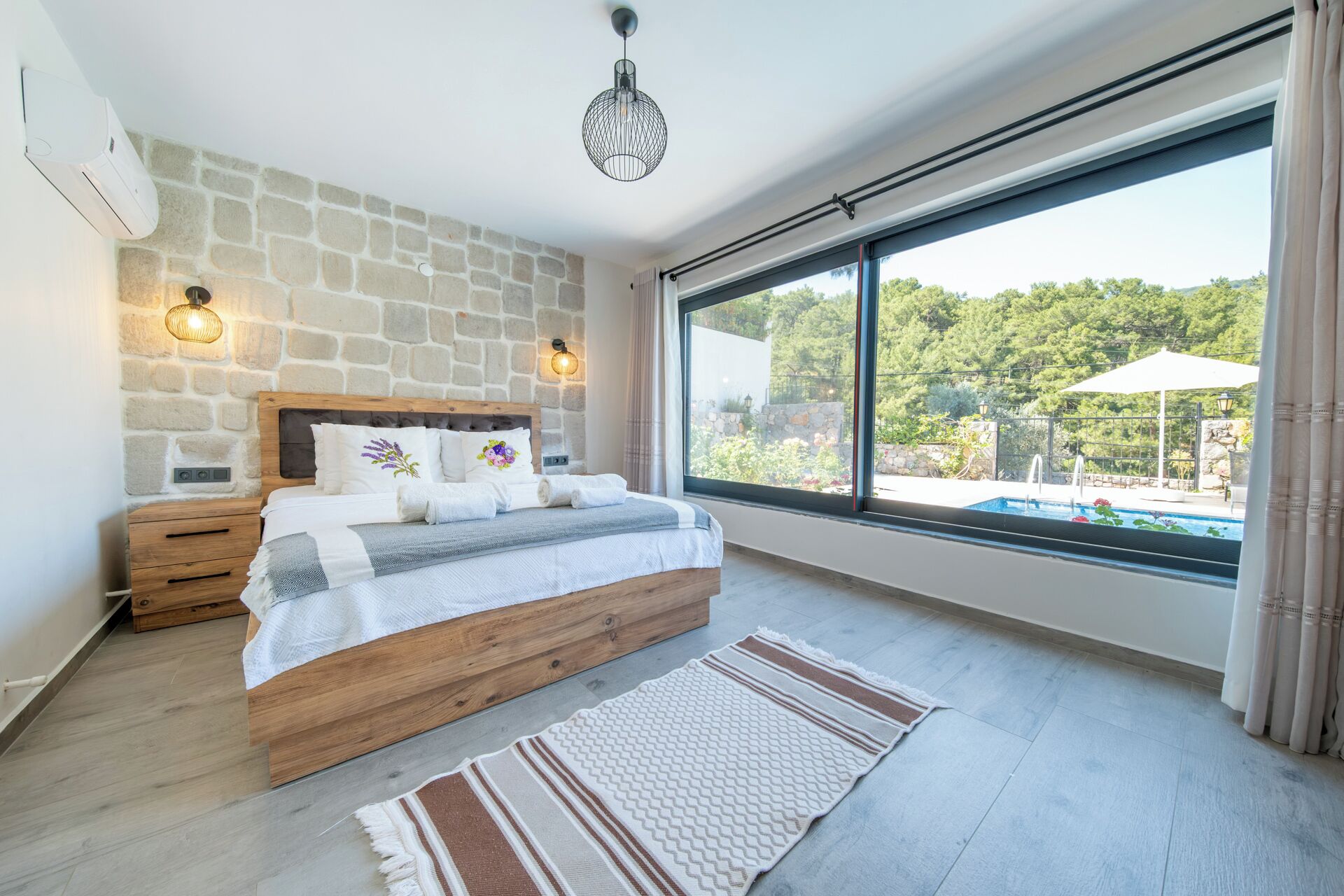 A bedroom with a stone wall, a wooden bed, a large window, and a view of a pool.