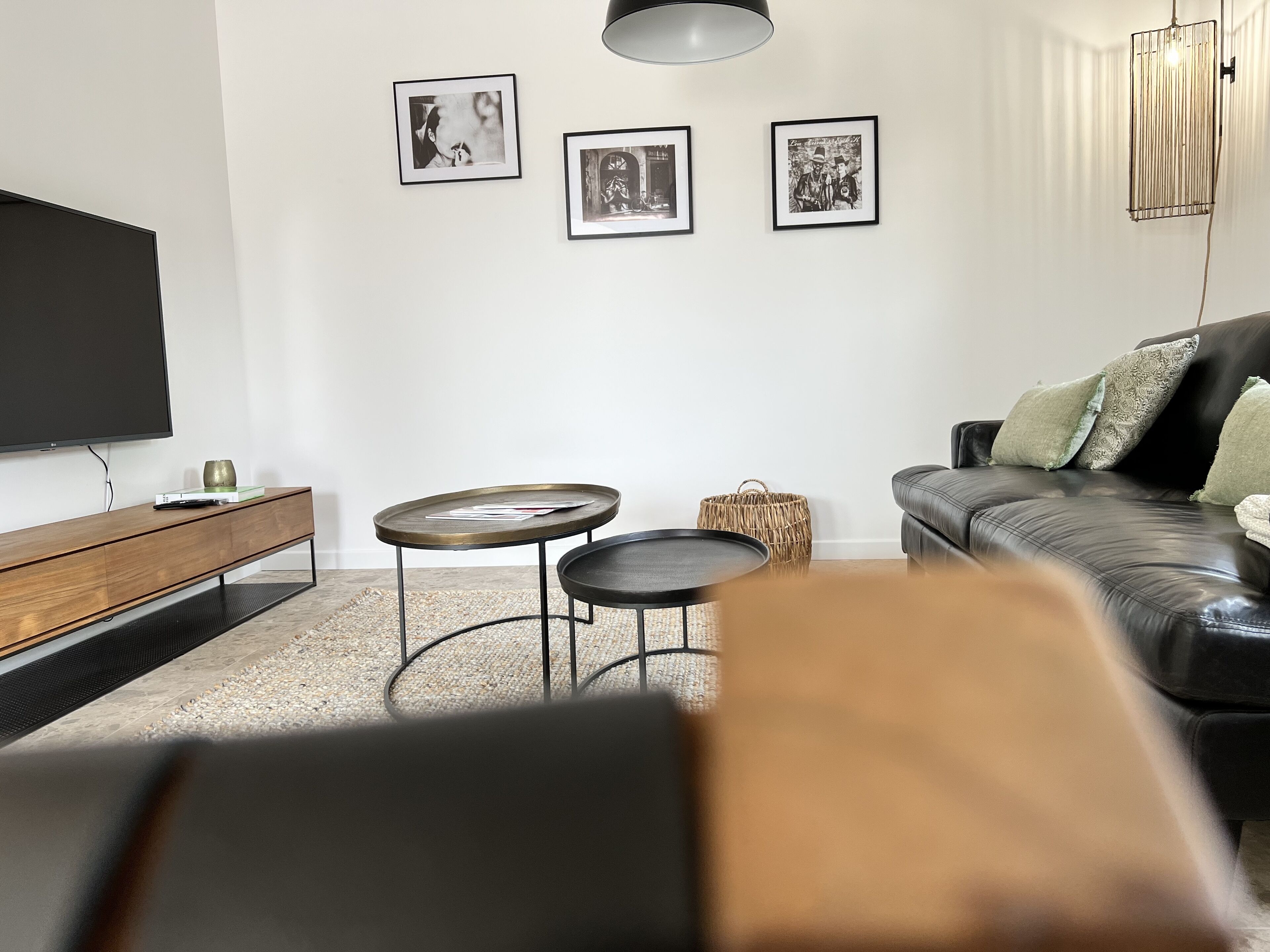 A modern living room with a flat-screen TV, a wooden console, two round tables, a black leather sofa, and three framed pictures on the wall.