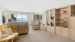 A spacious living room with a sofa, armchair, and a glass coffee table. There is a large cabinet with glass doors and a view of the outdoors through the windows. - Soldiers Point Absolute Waterfront The Peninsula (Soldiers Point)