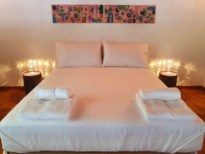 Ein ordentlich bezogenes Bett mit weißen Leinen und zwei Kissen, an dessen Seiten je ein Nachttisch mit Lampe steht, in einem Zimmer mit schlichter Wand und einem farbenfrohen, abstrakten Gemälde über dem Bett.