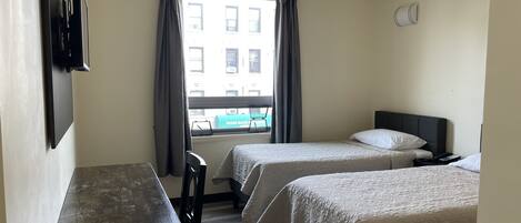 Habitación de hotel con dos camas, un escritorio, una silla, un ventanal con cortinas y vistas a edificios.